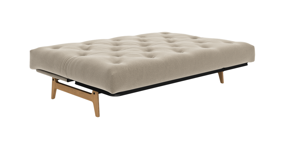 Beige udslået sovesofa/futon med tuftet madras og ben i lys eg, frilagt på hvid baggrund.