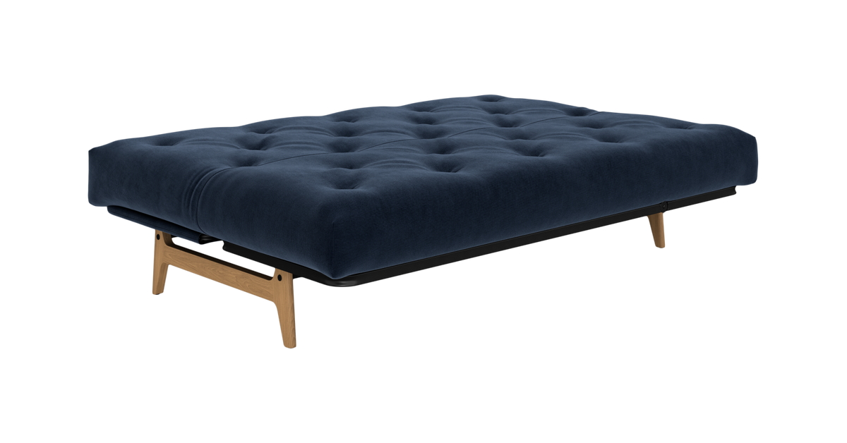 Mørkeblå, tuftet sovesofa/futon med træben og sort ramme, fotograferet skråt forfra på neutral baggrund.