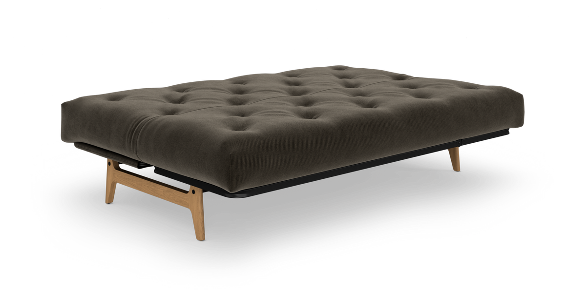 Fritlagt futon/sofaseng med tuftet mørkegrå madras og lyse egetræsben på hvid baggrund.