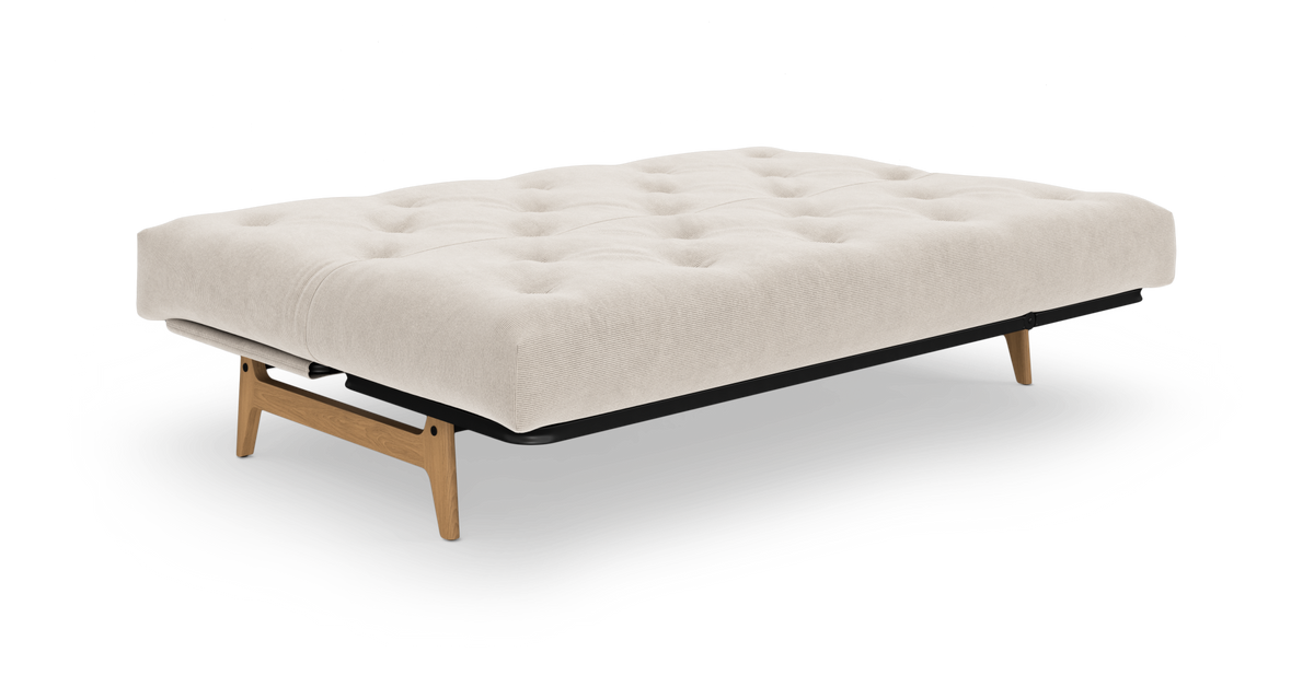 Beige futon/bæddemodul med tuftet madras, sort ram og lyse træben på hvid baggrund.