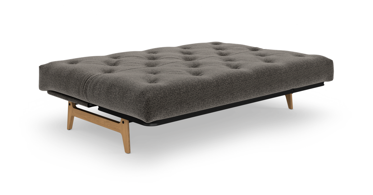 Grå tuftet sovesofa/futon med træben, frilagt på hvid baggrund.