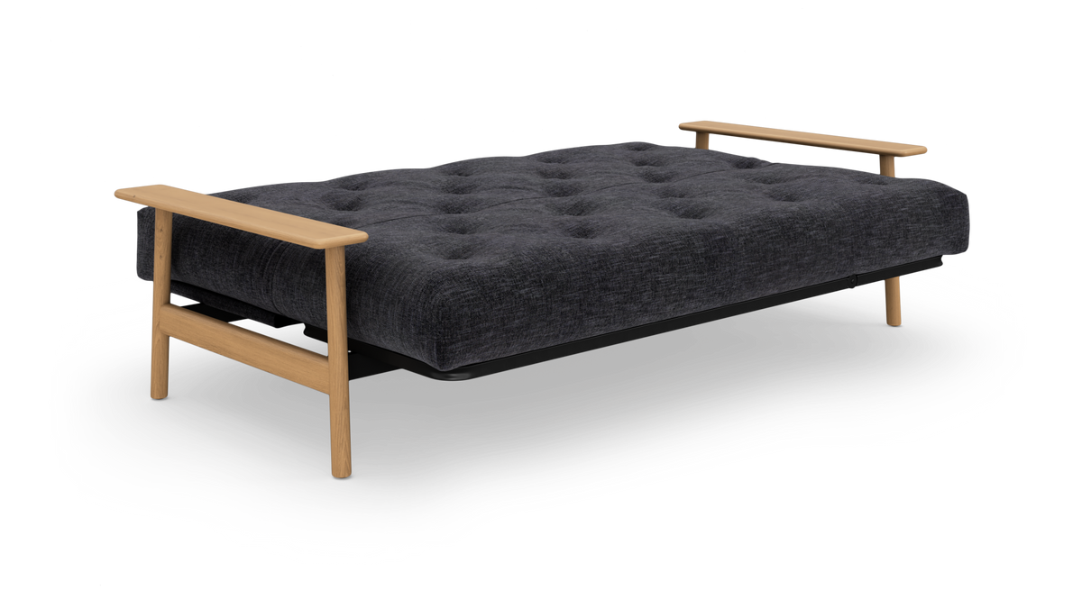 Mørkegrå sovesofa med tuftet madras, træarmlæn og ben, frilagt på hvid baggrund, set skråt forfra.
