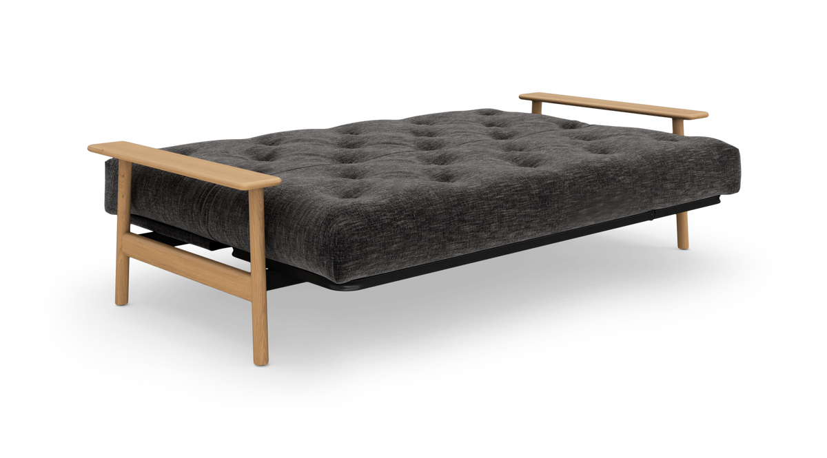 Mørkegrå sovesofa med knapquiltet madras og lyse træarmlæn, frilagt på hvid baggrund.