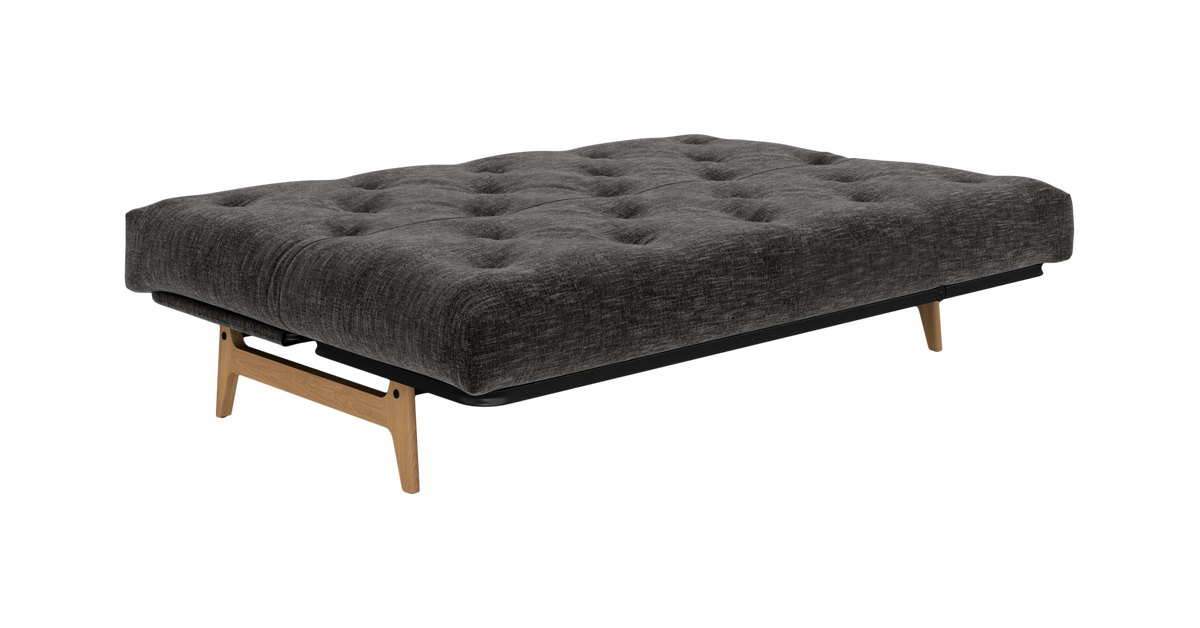Grå tufted futon/sovesofa med træben, frilagt på hvid baggrund.
