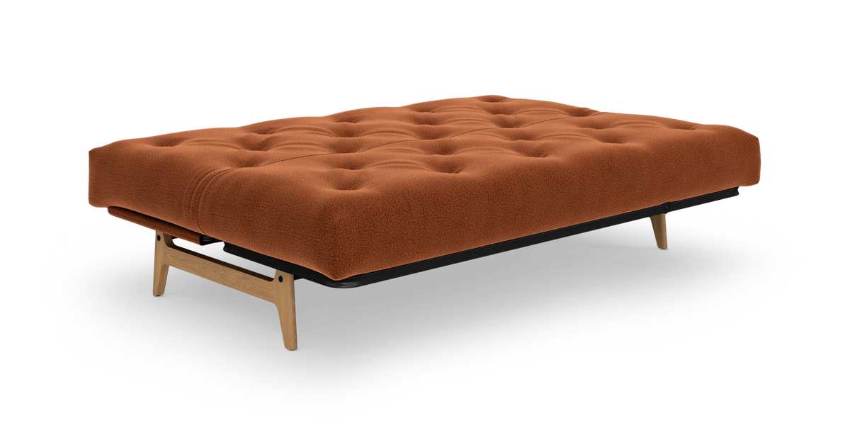 Lav, tuftet sovesofa/futon i rustfarvet tekstil med træben, frilagt på hvid baggrund.