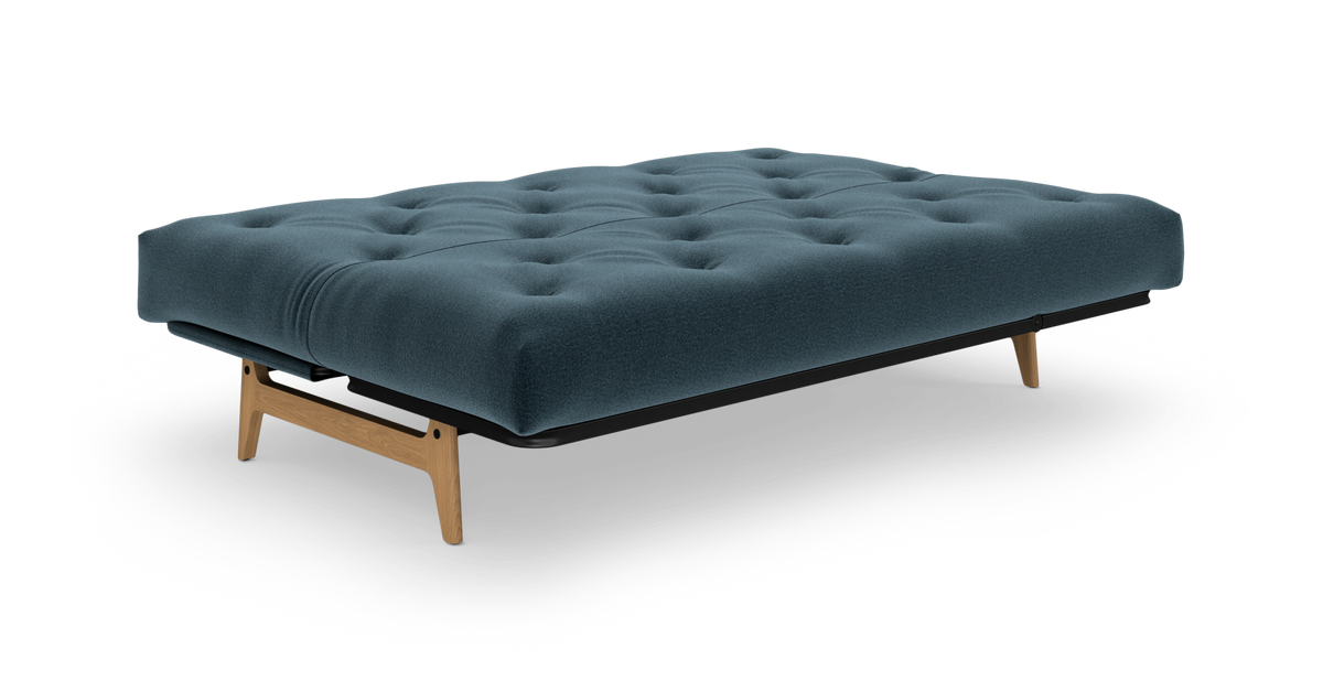 Blå futon/dagseng med tuftet madras og træben, frilagt på hvid baggrund.