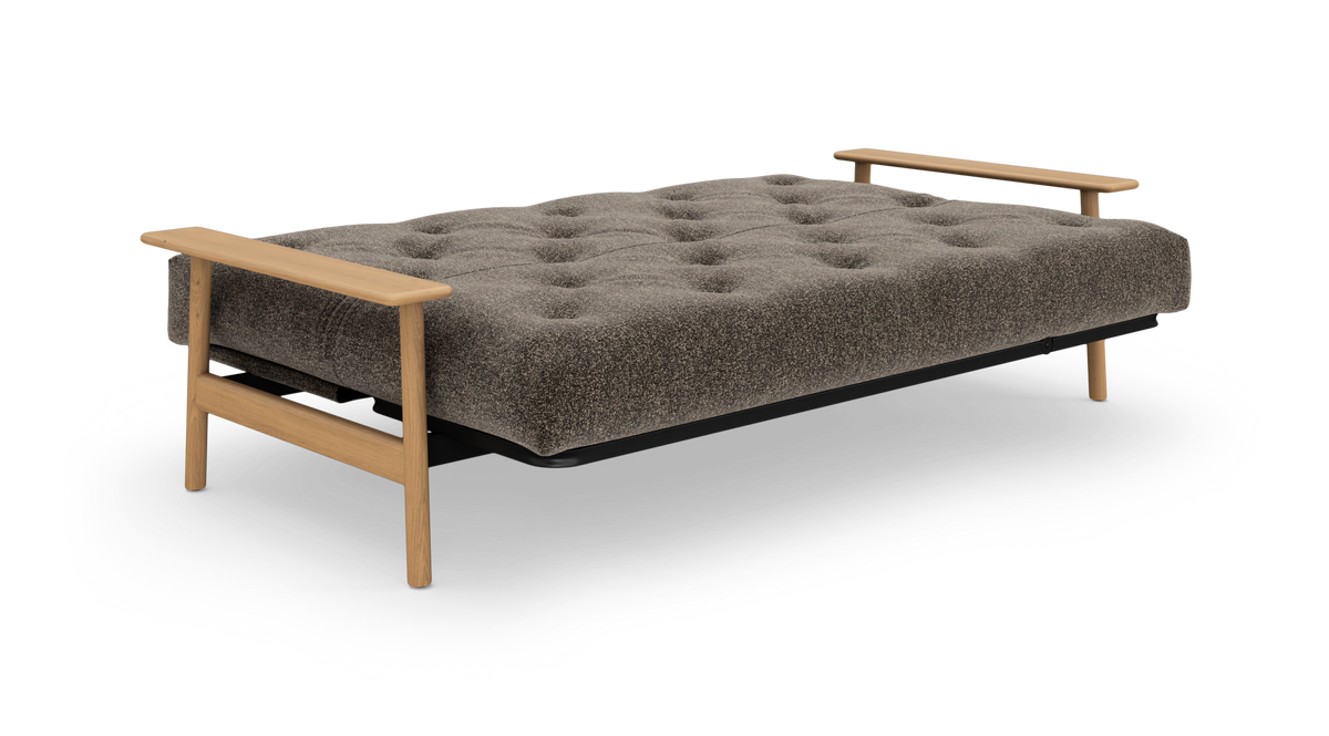 Bred sovesofa i udslået position med tuftet gråbrun madras og lyse træarmlæn på begge sider, frilagt på hvid baggrund.