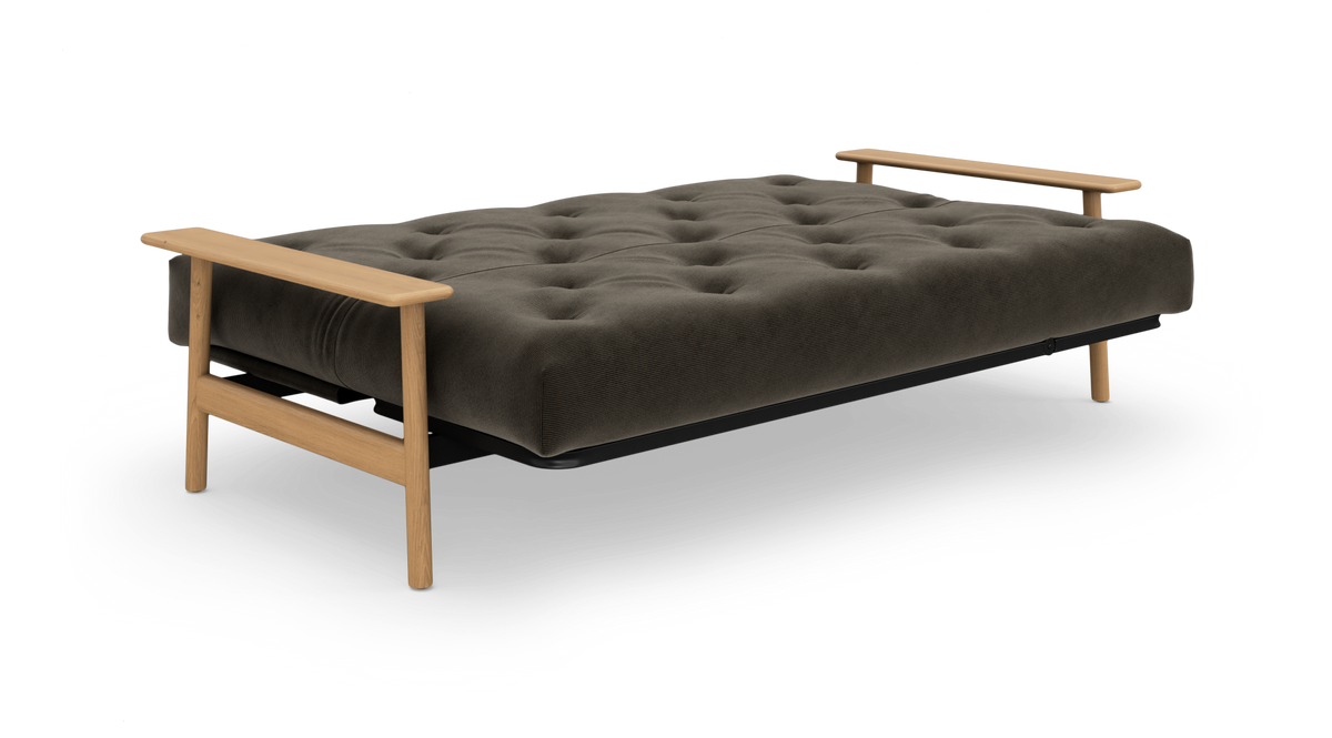 Mørkegrå sovesofa med knapquiltet madras og lyse træarmlæn og ben, frilagt på hvid baggrund set skråt forfra.