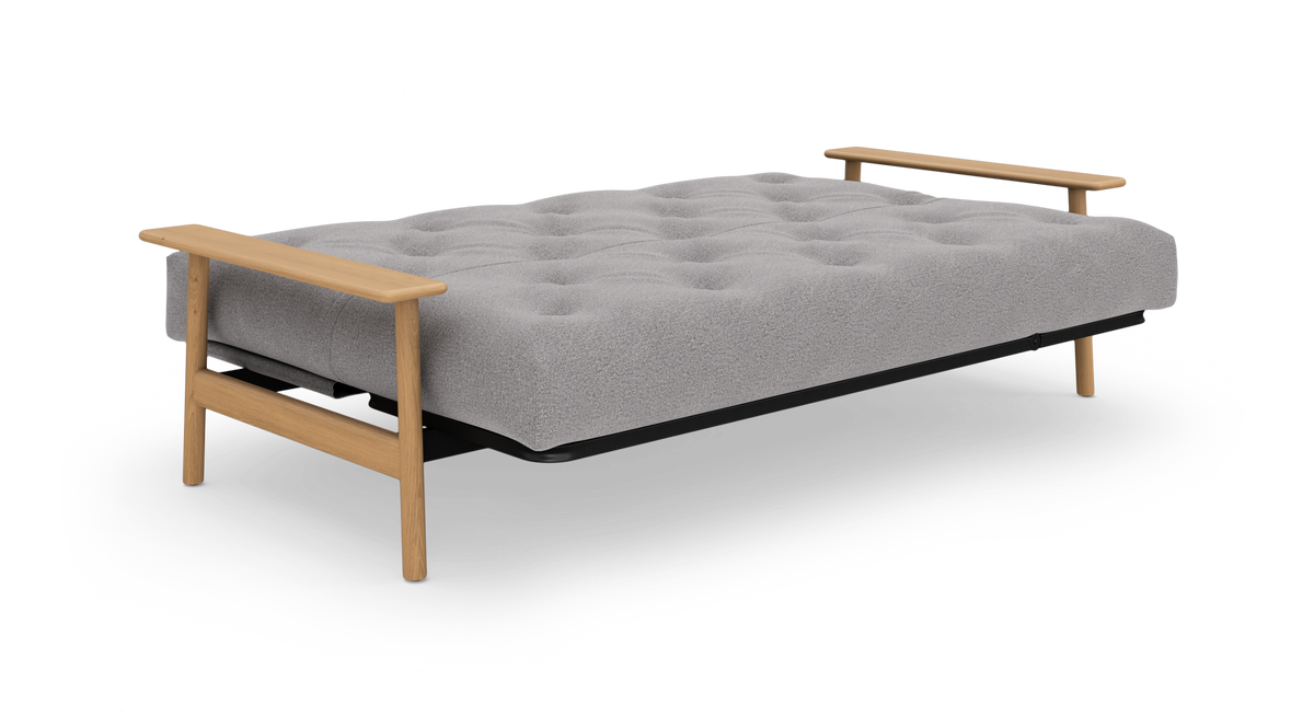 Grå sovesofa/futon med tuftet madras og lyse træarmlæn, frilagt på hvid baggrund.