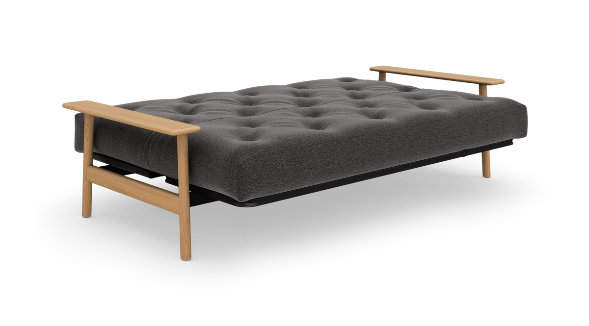 Grå sovesofa med tuftet madras og lyse træarmlæn, frilagt på hvid baggrund.