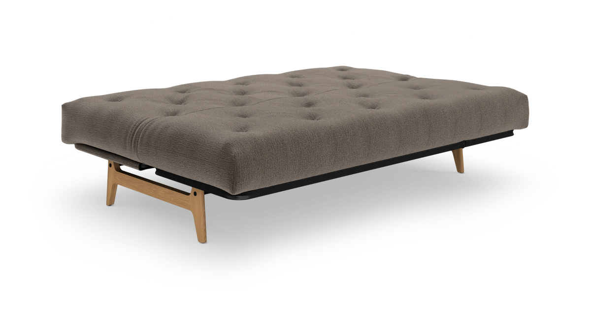 Lav sovesofa/daybed i beigegrå med knaptuftet madras, sort ram og ben i lys eg, frilagt på hvid baggrund.