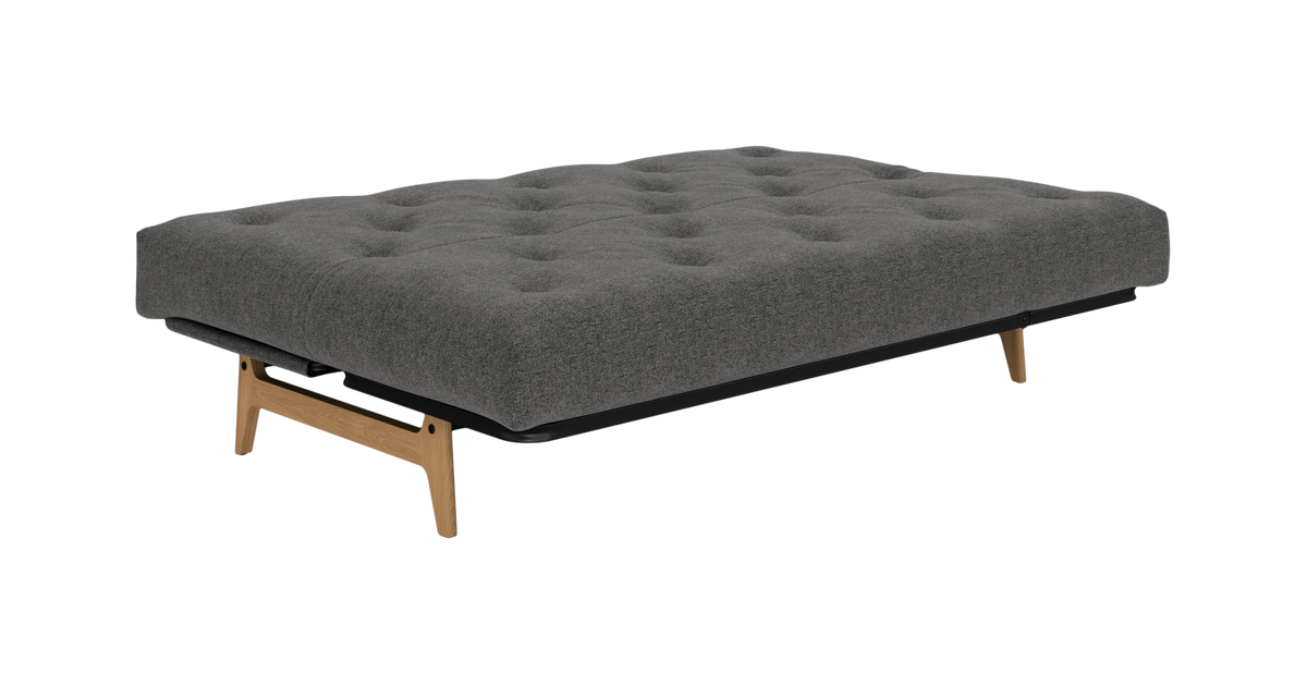 Grå sovesofa/futon med tuftet madras og træben, fritlagt på hvid baggrund.