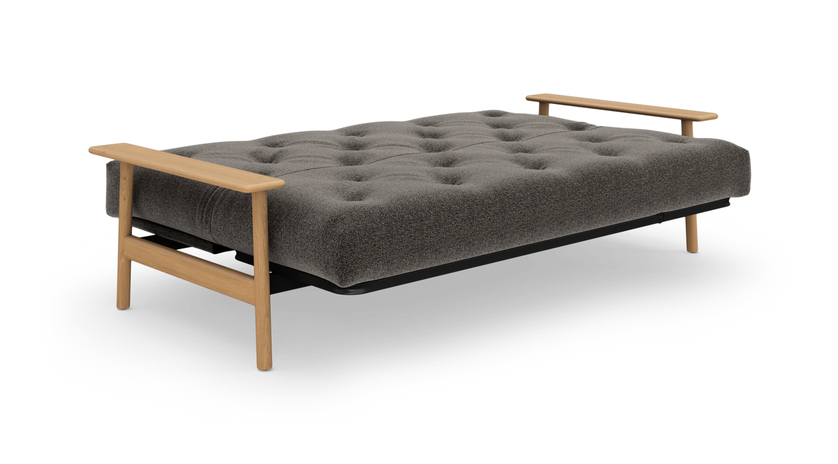 Grå, tufted sovesofa/futon med armlæn i egetræstoner, frilagt på hvid baggrund.