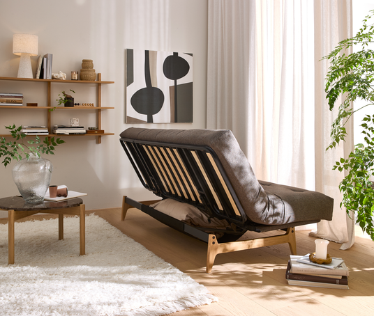 Lyst skandinavisk stue med sovesofa/futon i side, løftet ryg, treramme, blød tæppe, væghylder med lampe og kunst, grønne planter og lyse gardiner i blødt dagslys.