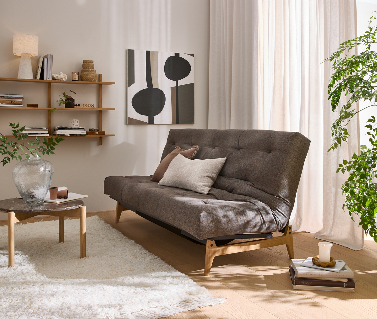 Lys skandinavisk stue med brun sovesofa på træben, hvide gardiner, væghylde med lampe og dekoration, grøn plante, hvid shaggy tæppe og lille sofabord i træ/marmor.