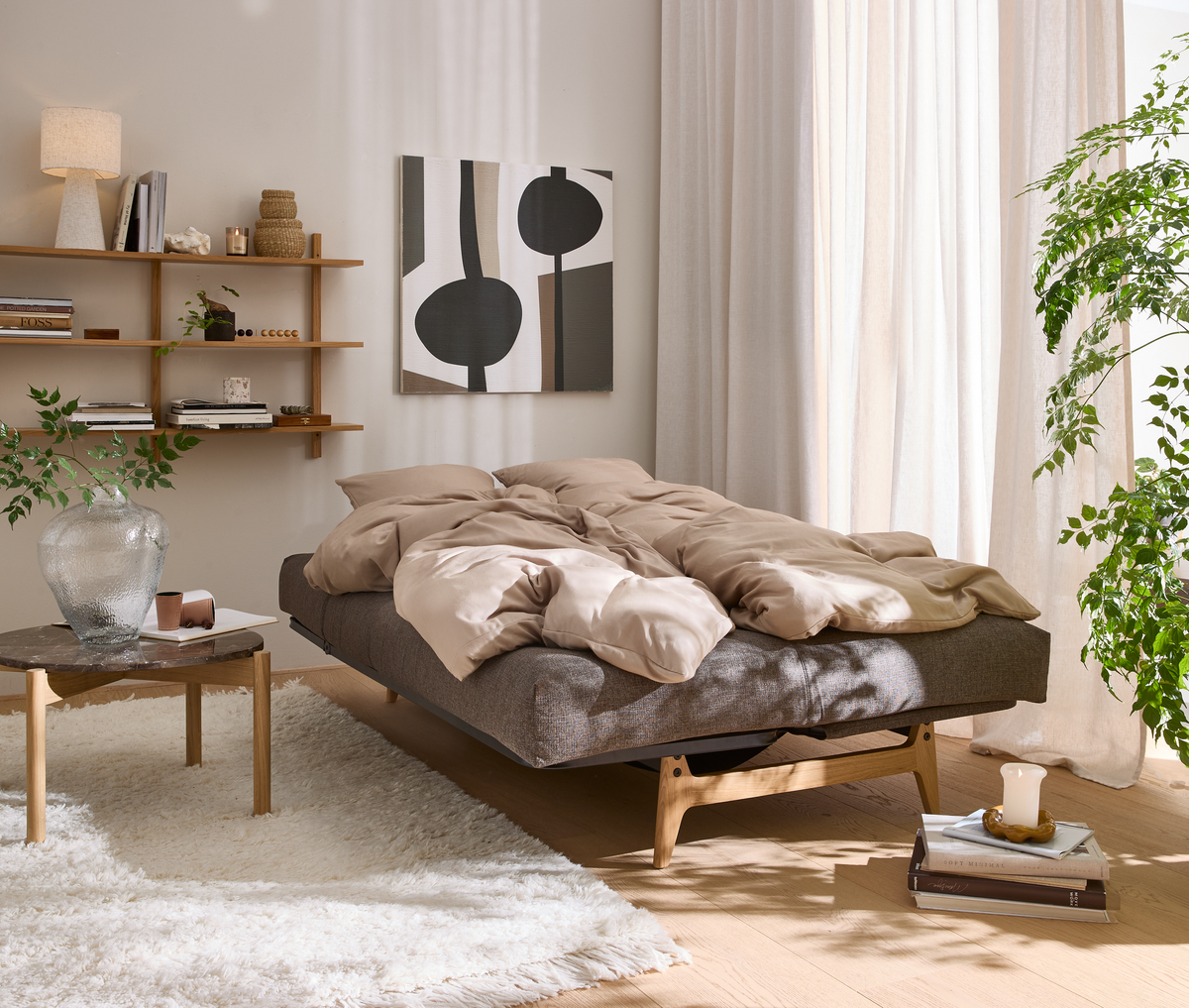 Lyst og roligt soveværelse med grå seng på træben, beige sengetøj, vægkunst, hylde med bøger og lampe, grønne planter samt lyse gardiner i dagslys.