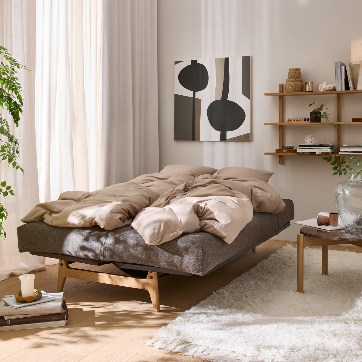 Lys skandinavisk stue med grå daybed, beige sengetøj, blødt hvidt tæppe, træhylde og planter; morgensol gennem store gardiner.