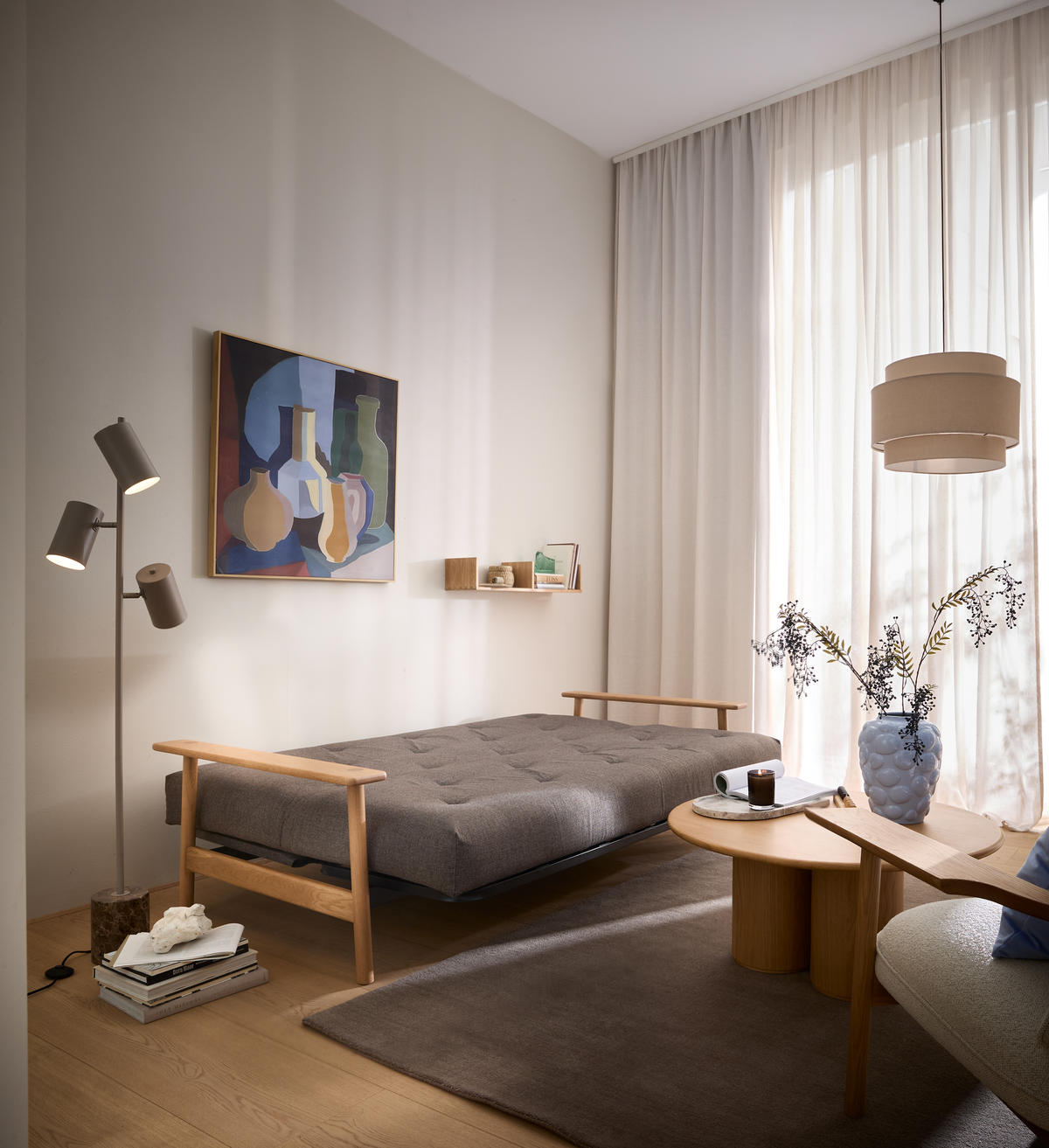 Lyst skandinavisk stue med grå daybed, runde træborde, gulvlampe, abstrakt vægkunst og lyse gardiner i blødt dagslys.