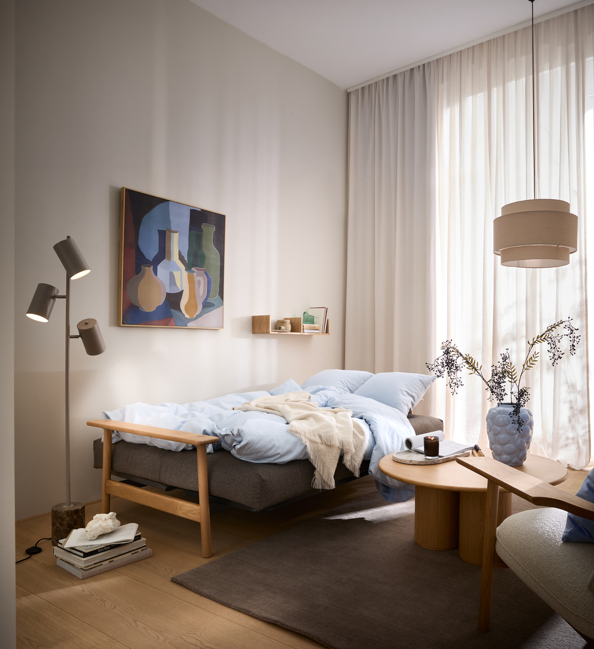 Lyst, skandinavisk stue med daybed med lyseblåt sengetøj, træborde, gulvlampe, abstrakt vægkunst og lyse gardiner i dagslys.