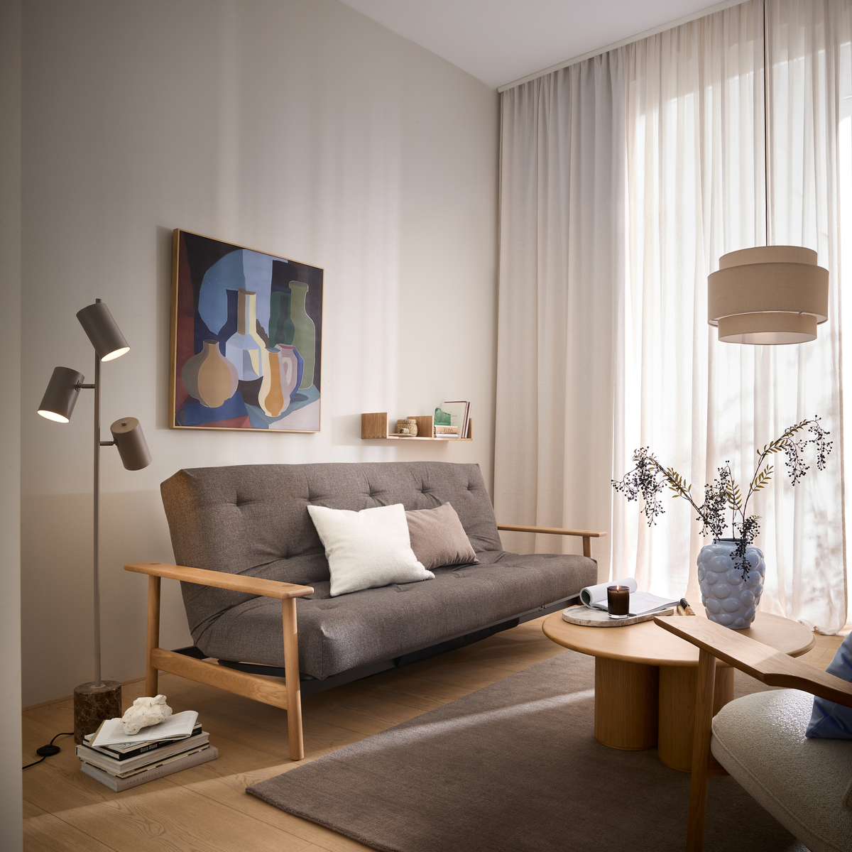 Lyst skandinavisk stue med grå sofa, trædetaljer, standerlampe, abstrakt vægkunst, runde sofaborde med vase og tændt lys, store lyse gardiner og bløde neutrale nuancer.