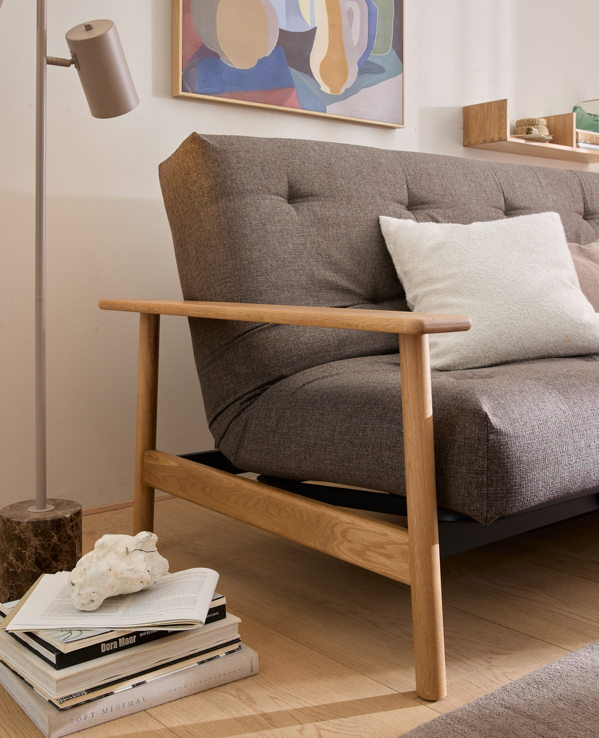Nærbillede af brun sofa med træarmlæn og hvid pude, gulvlampe med stenfod og en bogstak på gulvet med titlerne Dora Maar og Soft Minimal; abstrakt kunst på væggen.