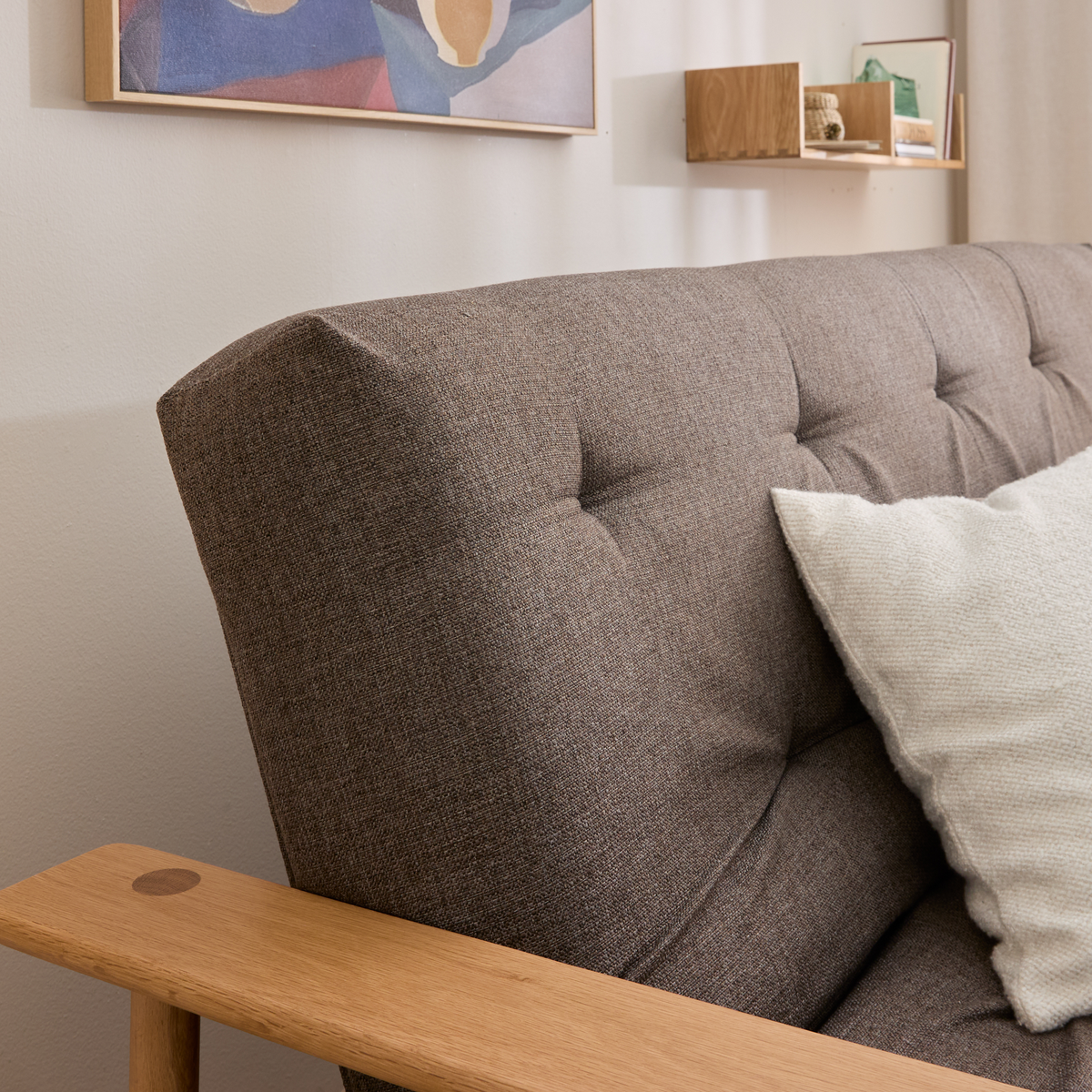 Nærbillede af brun stofsofa med knap-quiltet ryg, lyst træarmlæn og cremefarvet pude; væg med kunst og lille hylde i baggrunden.