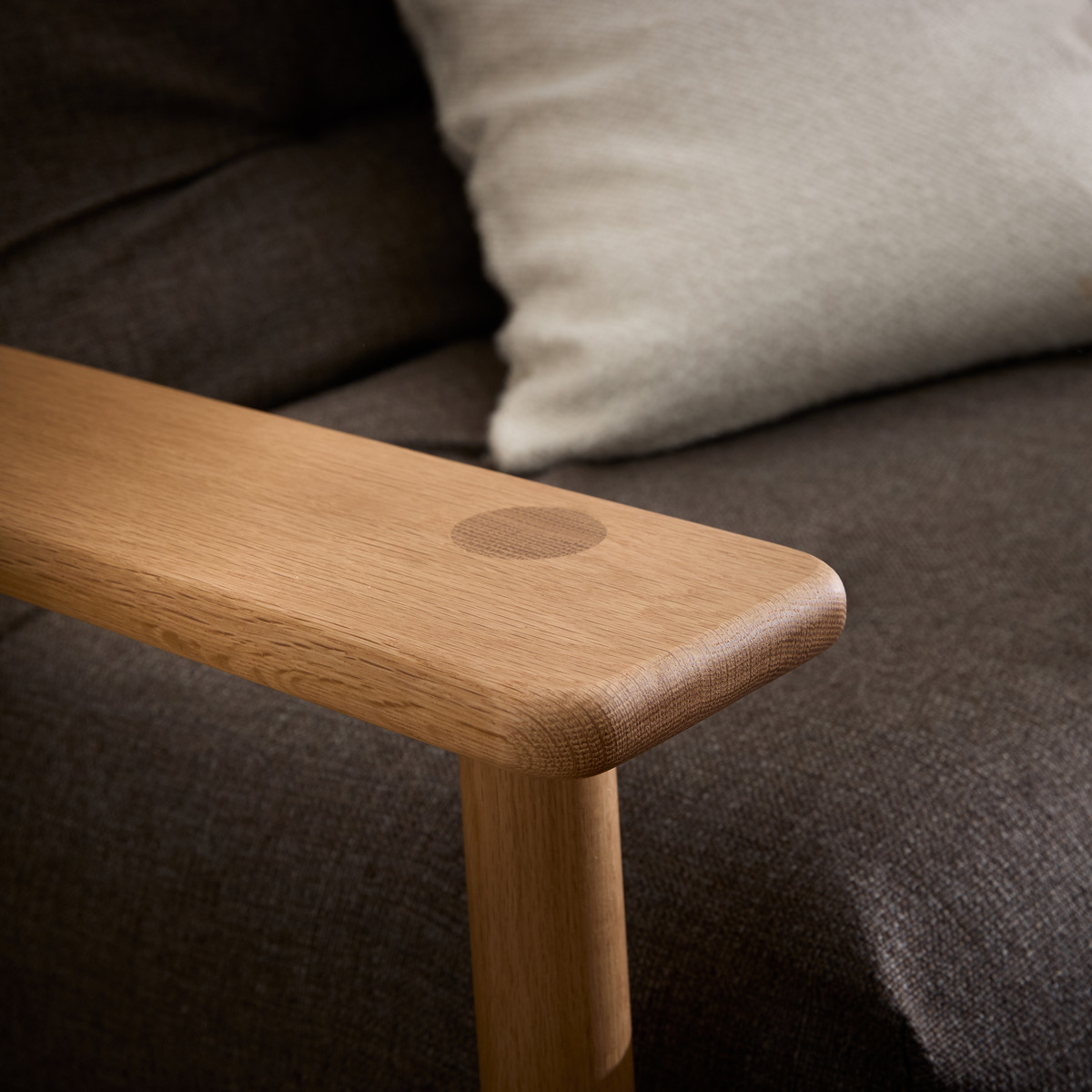 Nærbillede af et armlæn i lys eg med afrundede kanter på en brun stofsofa med en lys beige pude.