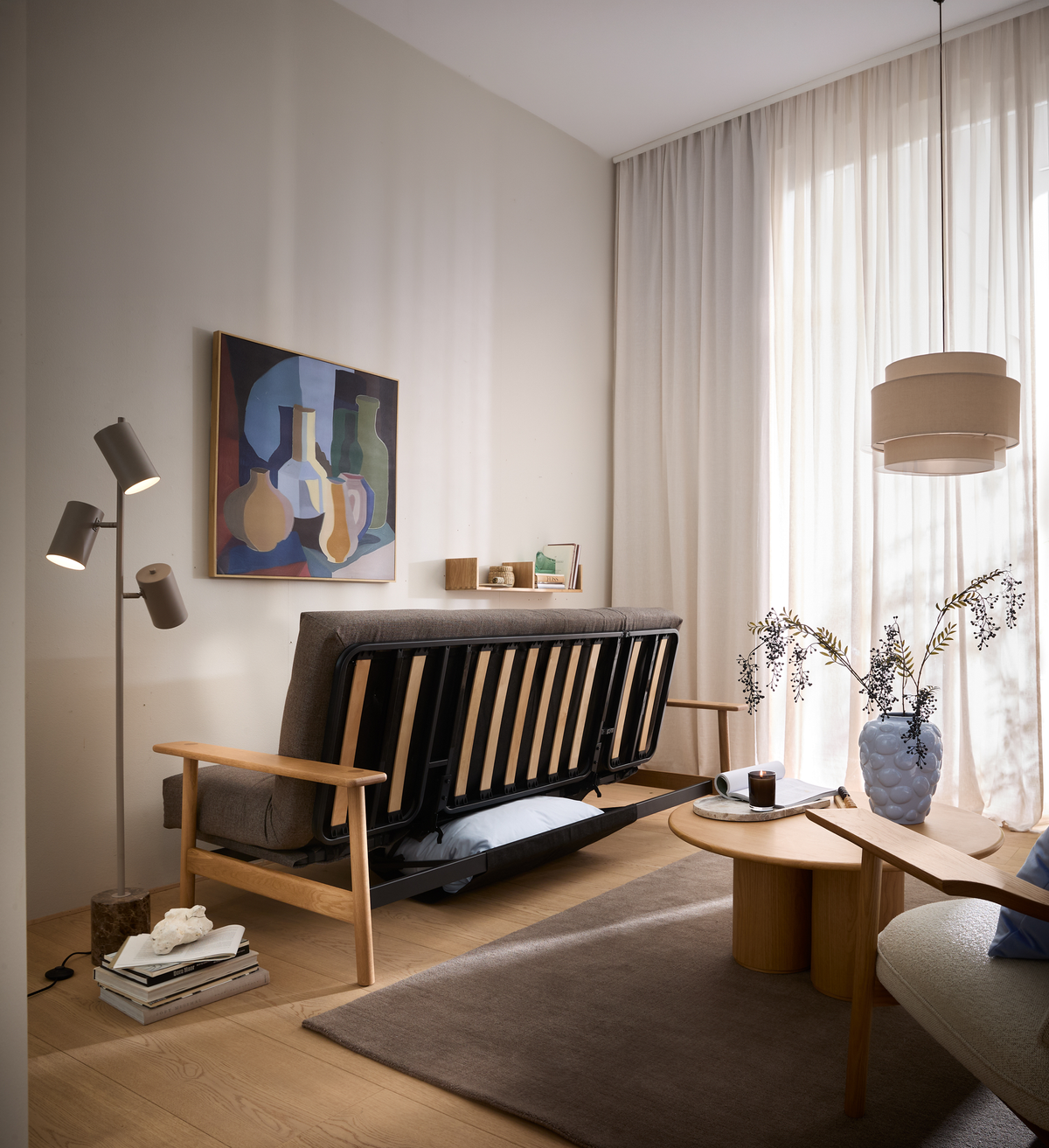 Skandinavisk stue i lyse beige nuancer med en sovesofa hvor senge-rammen er slået op, lette gardiner, træ-sofabord, gulvlampe, pendel og en vase med grene på bordet.