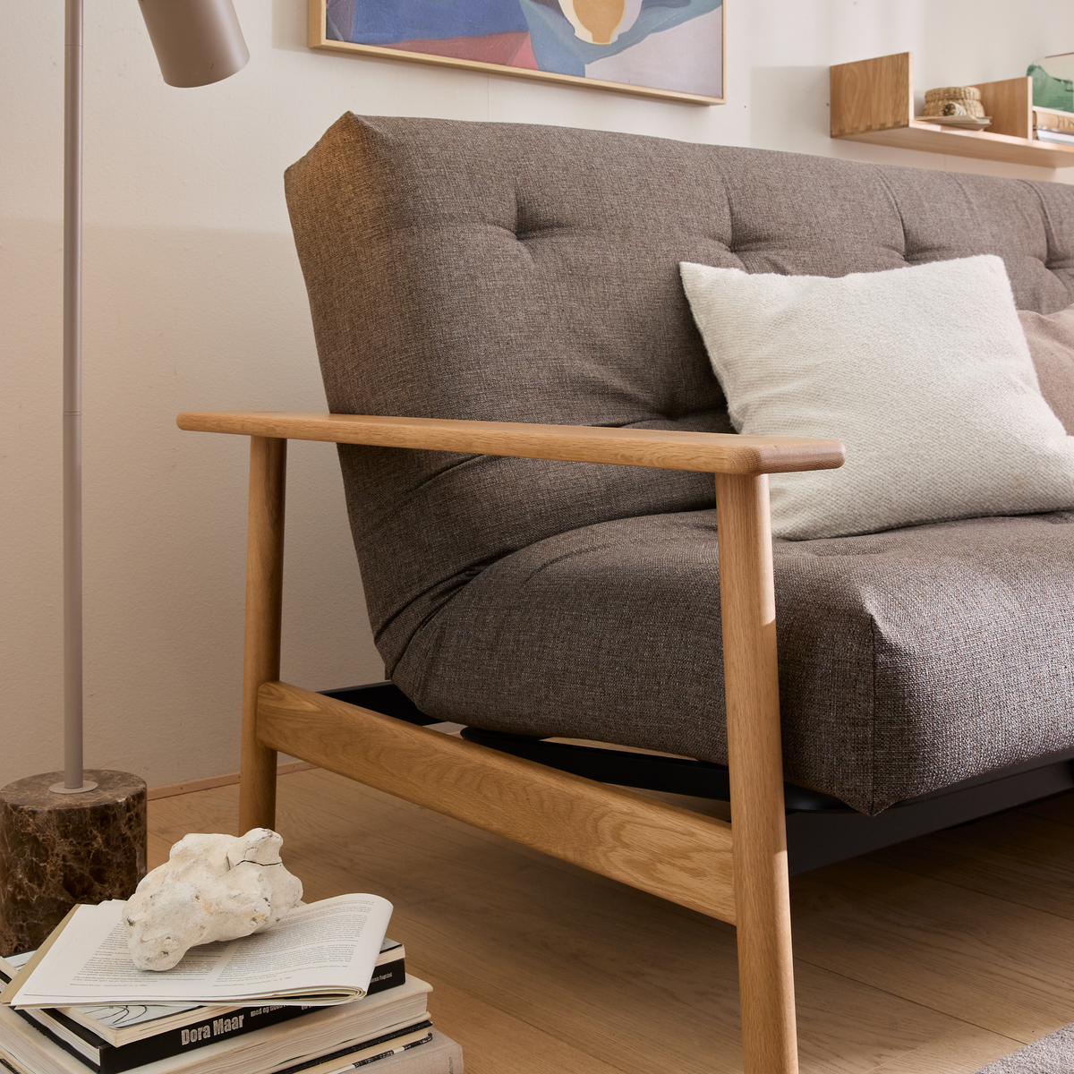 Nærbillede af brun sofa med træarmlæn og hvid pude; gulvlampe, vægkunst og en bogstak med Bora Maar i en lys stue.