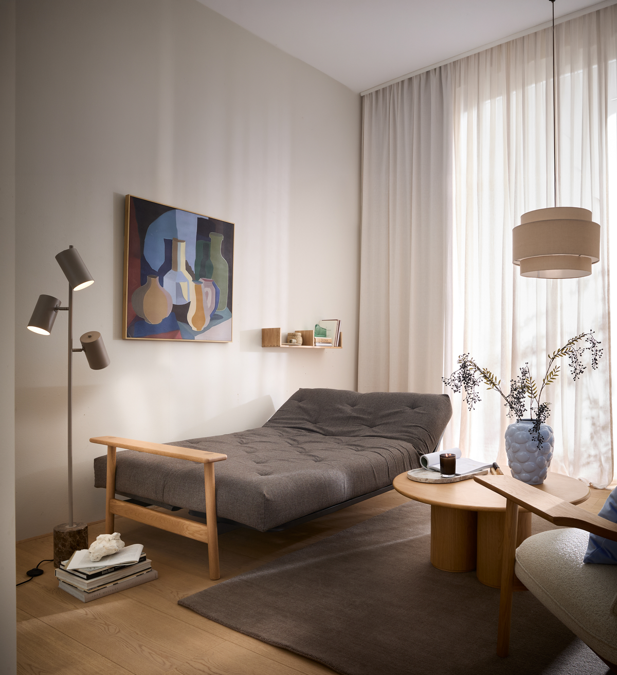 Lyst, skandinavisk stuehjørne med grå daybed, træborde, blå vase med grene, gulvlampe og abstrakt vægbillede; lette gardiner og naturligt dagslys.