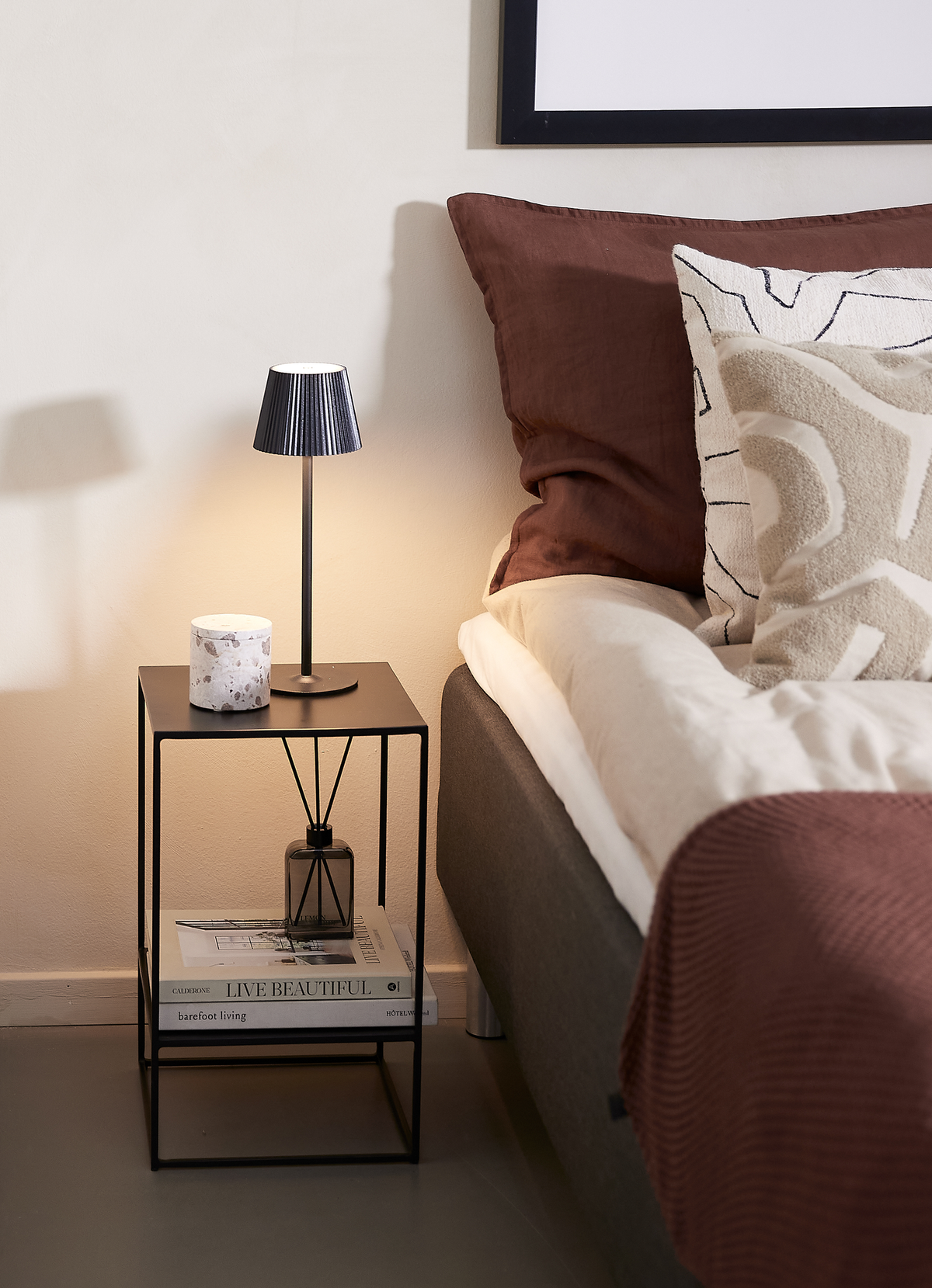 Hyggelig sengebordsscene: et sort sidebord med lille bordlampe, marmorlignende lys, duftpinde og bøgerne “Live Beautiful”, “barefoot living” og “Hotel” ved siden af en seng med rustbrunt og cremefarvet sengetøj.
