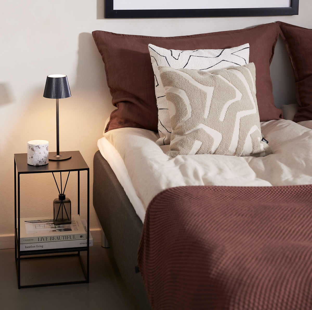 Sengehjørne med beige dyne, brun plaid og puder i beige og hvid; sort sengebord med lille sort bordlampe, duftpinde og bøgerne LIVE BEAUTIFUL og barefoot living.