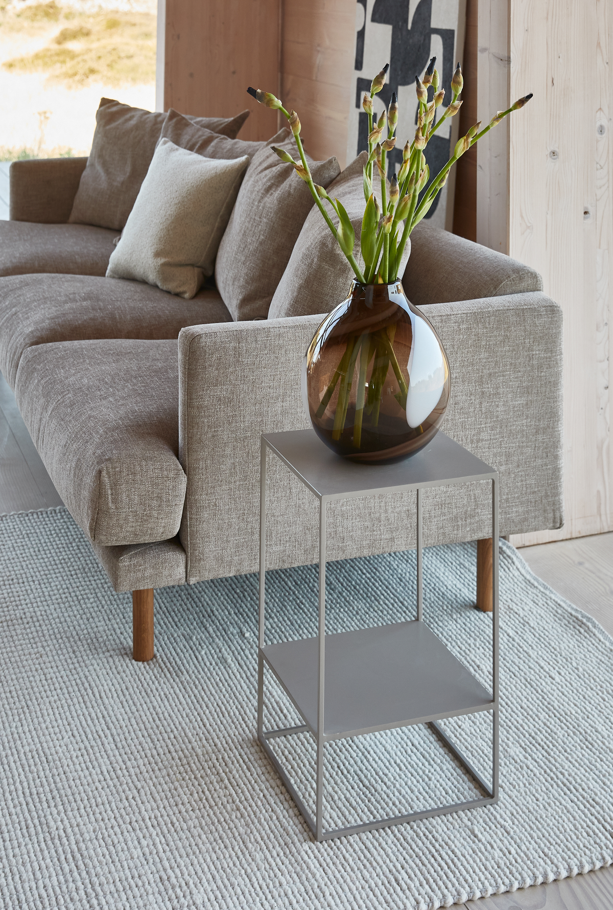 Beige sofa med puder, slankt metal-sidebord og brun glasvase med grønne knopper på lyst tæppe i en lys stue.