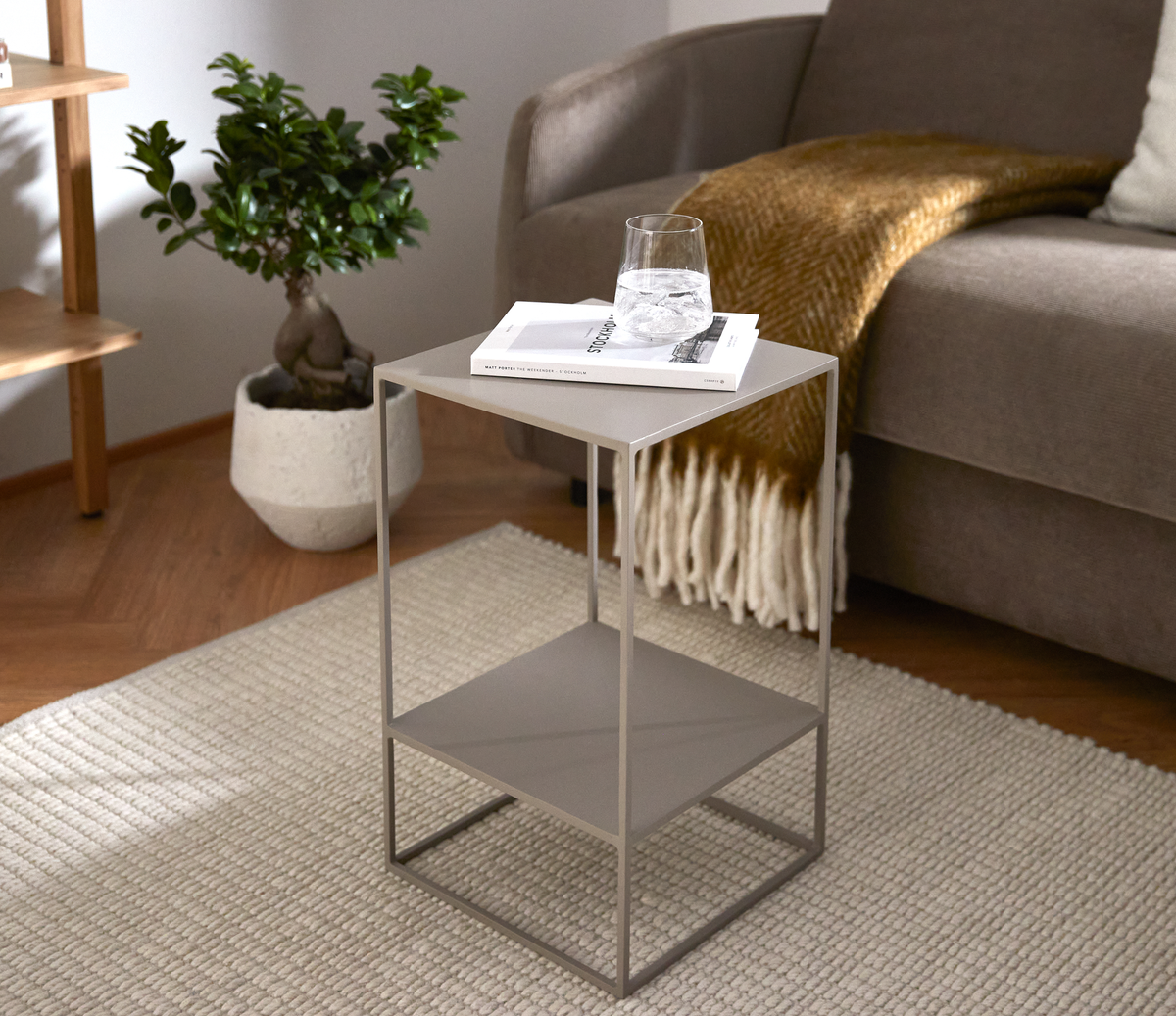 Moderne sidebord i metal med hylde, et glas vand og en bog ved siden af en sofa med sennepsgul plaid i en lys stue.