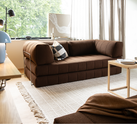 Brun modulsofa i en moderne stue med lyst, tekstureret tæppe, lille træbord og lette gardiner ved et stort vindue; sort‑hvid ternet pude på sofaen.