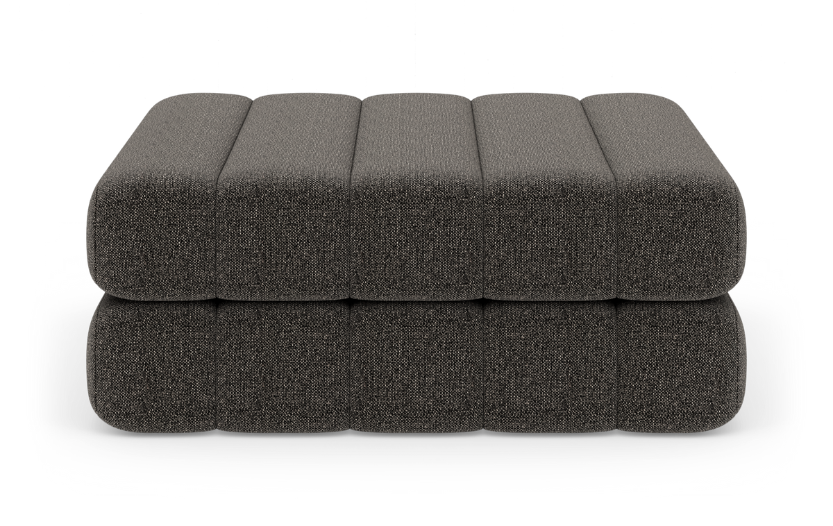 Mørkegrå polstret puf/ottoman i modulopbygget, tolags design med afrundede kanter, frilagt på hvid baggrund.