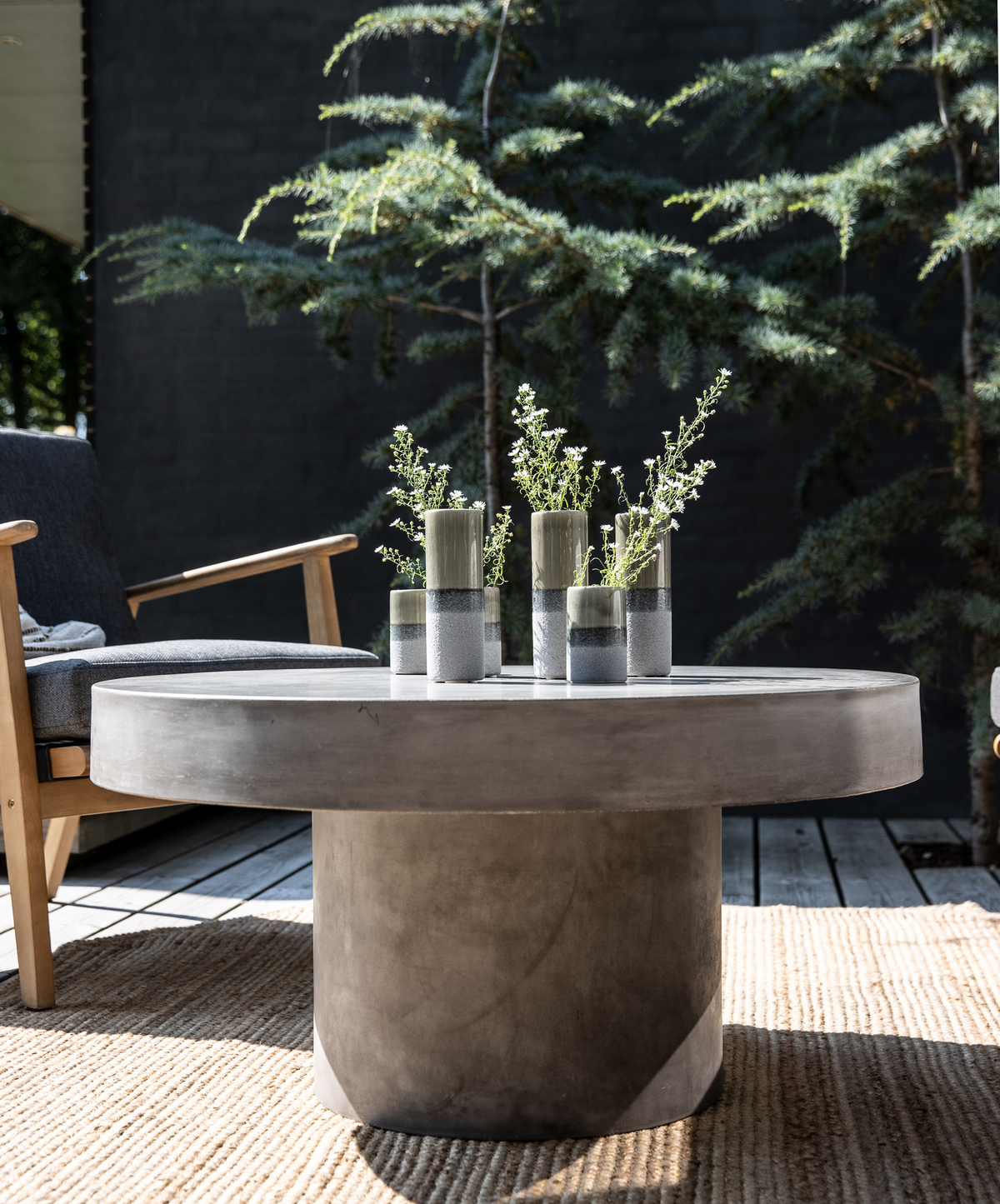 Rundt sofabord i betonlook på terrasse med små cylindervaser med grønne kviste på et jutetæppe ved siden af en loungestol.