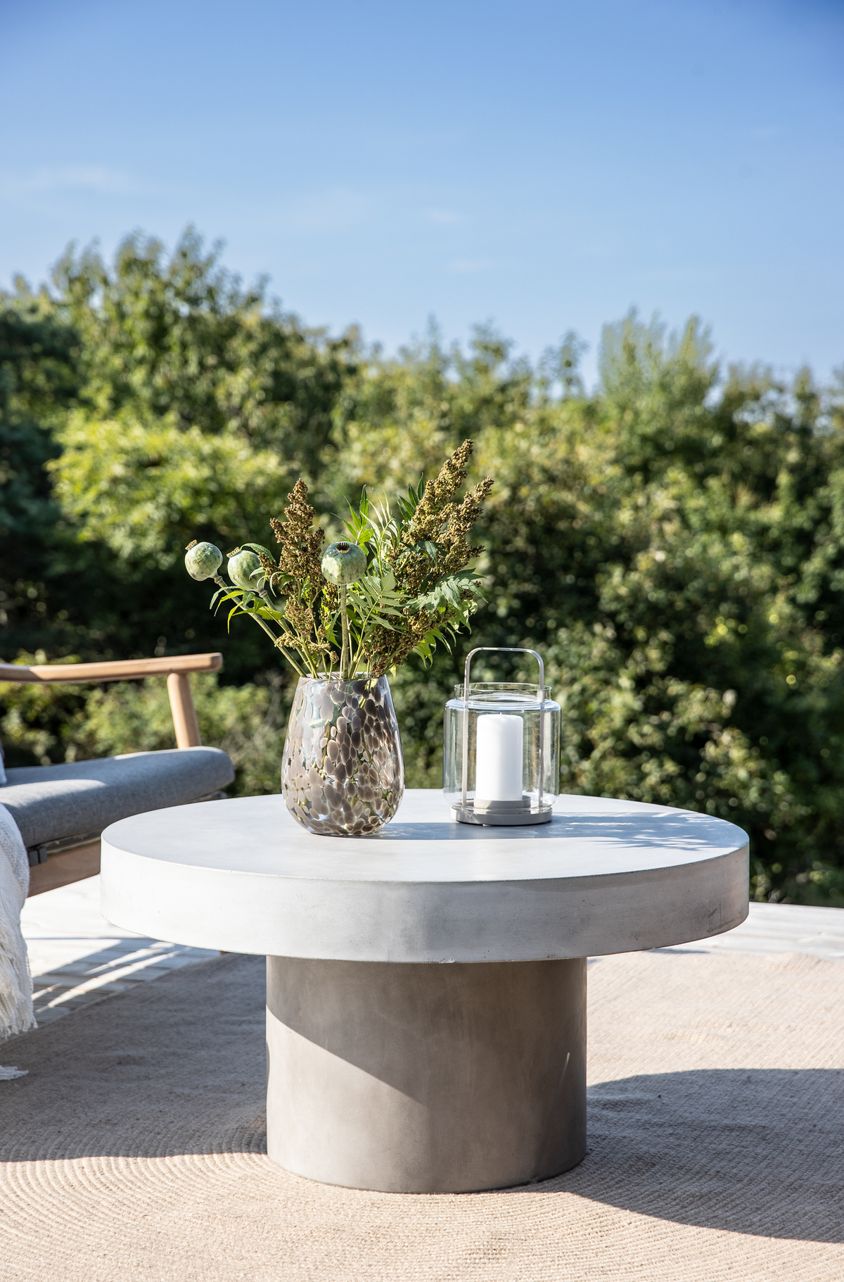 Rundt betonbord på terrasse med vase med grønne grene og glaslanterne med hvidt lys, solrig dag og grøn beplantning i baggrunden.