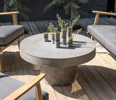 Rundt betonbord udendørs med små keramikvaser med grønne kviste på et jutetæppe mellem grå loungestole.