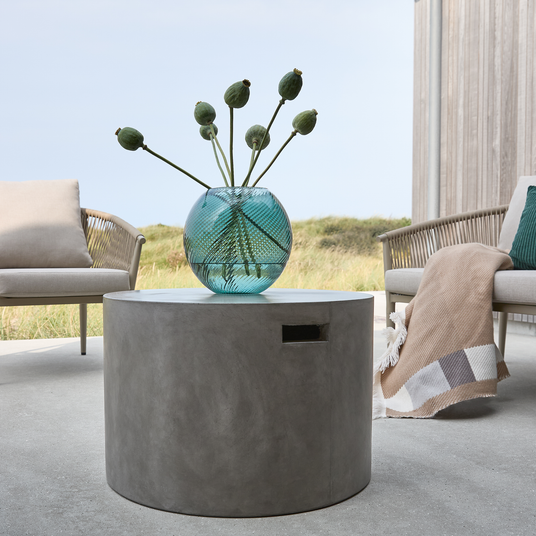 Rundt betonbord på terrasse med turkis glasvase og grønne frøkapsler, loungestole og plaid i baggrunden.