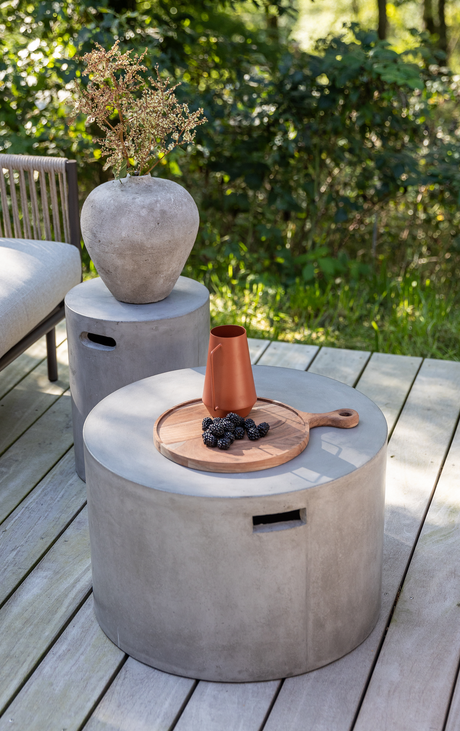 Rundt betonbord på træterrasse med træbakke, terrakottakande og brombær; ved siden af et cylindrisk sidebord med grå vase og tørrede grene i grøn udendørs omgivelser.