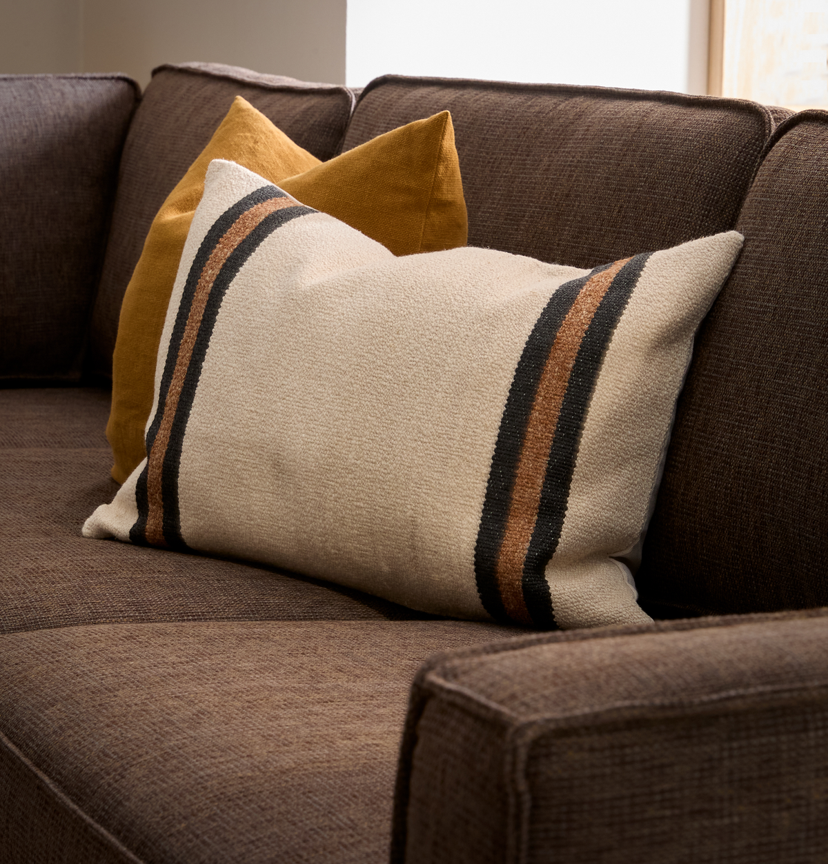 Nærbillede af brun sofa med to puder – en beige med sorte og brune striber og en sennepsgul – i blødt dagslys ved et vindue.