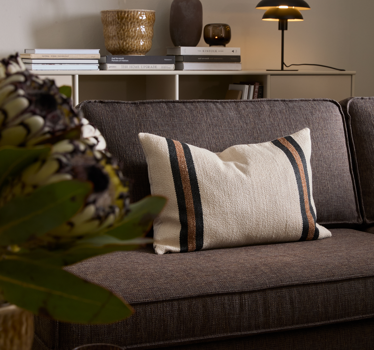 Brun sofa med beige pude med sorte og brune striber; i baggrunden en reol med bøger som Kinfolk og The Home Upgrade, keramik og en tænd bordlampe.