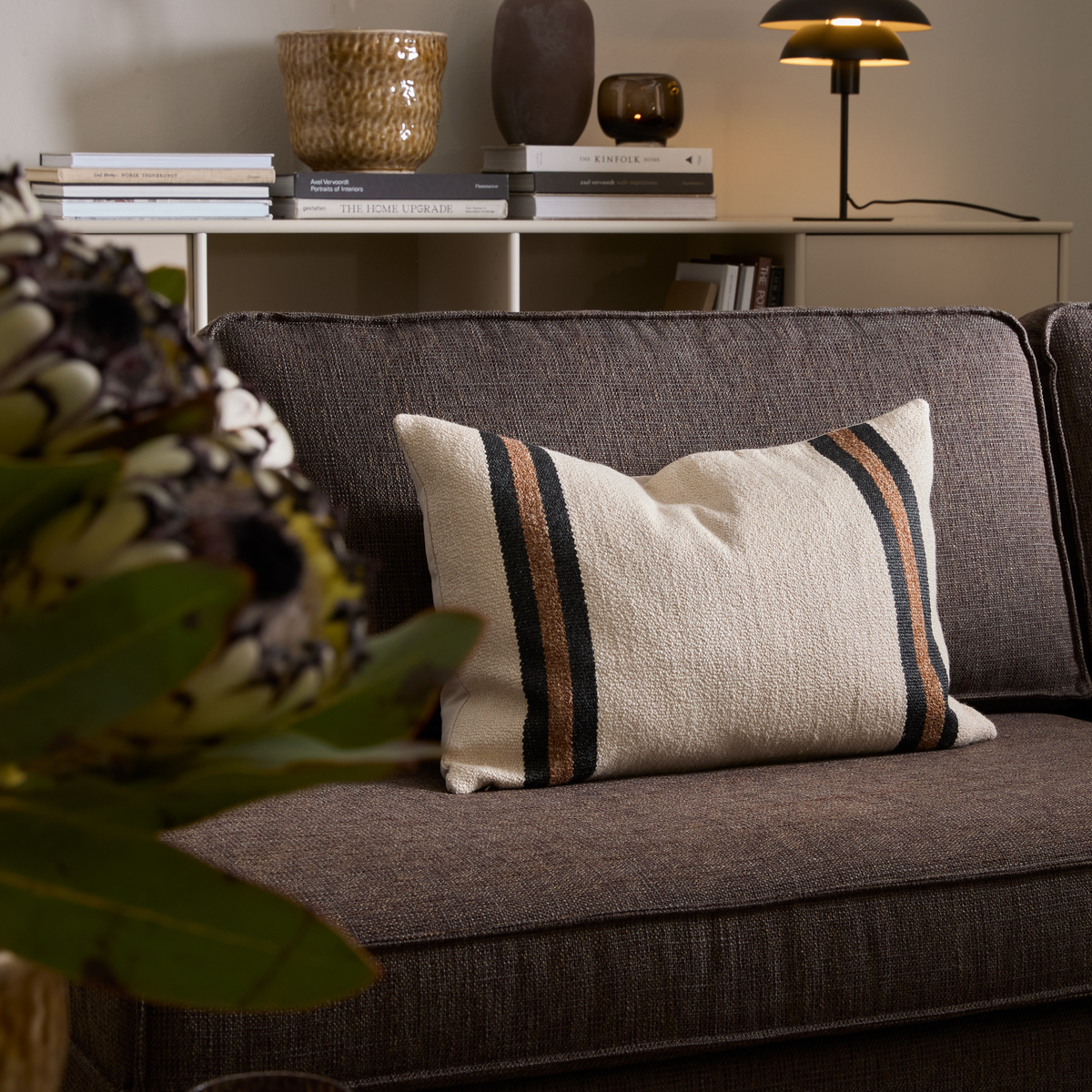 Beige rektangulær pude med sorte og brune striber på en brun sofa i en hyggelig stue med bordlampe og reol i baggrunden.