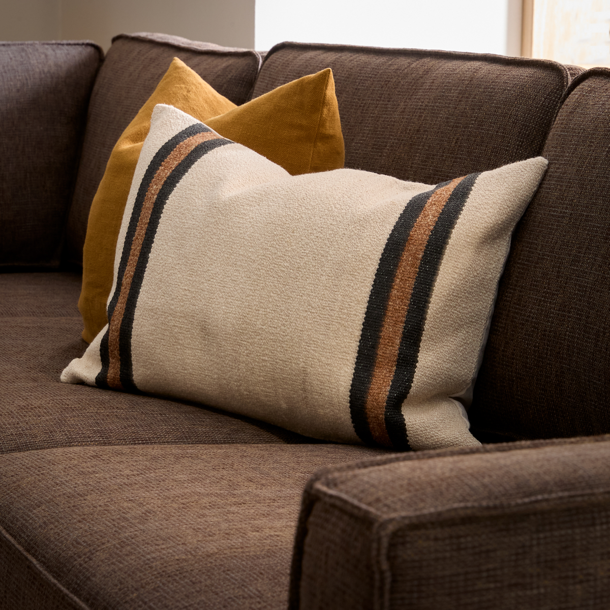 Brun sofa med beige pude med sorte og brune striber og en sennepsgul pude bagved i blødt dagslys.