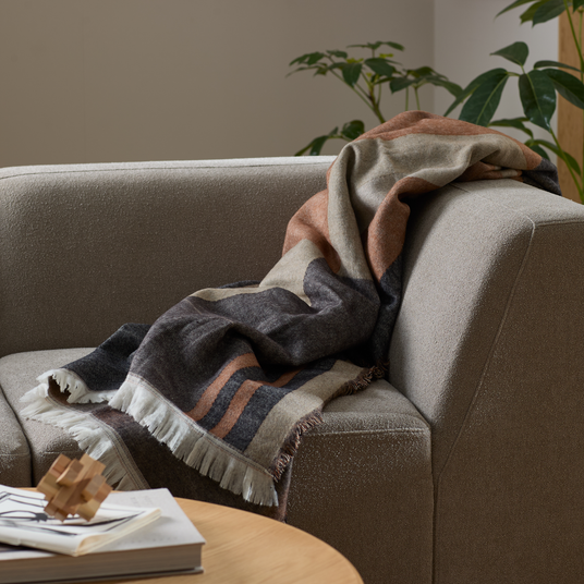 Vævet plaid med frynser lagt over armlænet på en beige sofa i stuen, med grønne potteplanter i baggrunden og bøger med et træpuslespil på sofabordet.