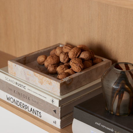 Stilleben på en træhylde: et stenfad fyldt med valnødder ligger oven på bøgerne “SCANDI RUSTIC”, “THE KINFOLK TABLE”, “WONDERPLANTS” og en sort bog med ryggen “axel vervoordt wabi”, ved siden af en lille stribet glasvase.
