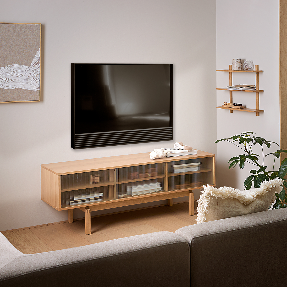 Lyst skandinavisk stue med vægmonteret TV over et lavt mediemøbel i lyst egetræ, grå sofa, lille hylde og grøn potteplante i dagslys.