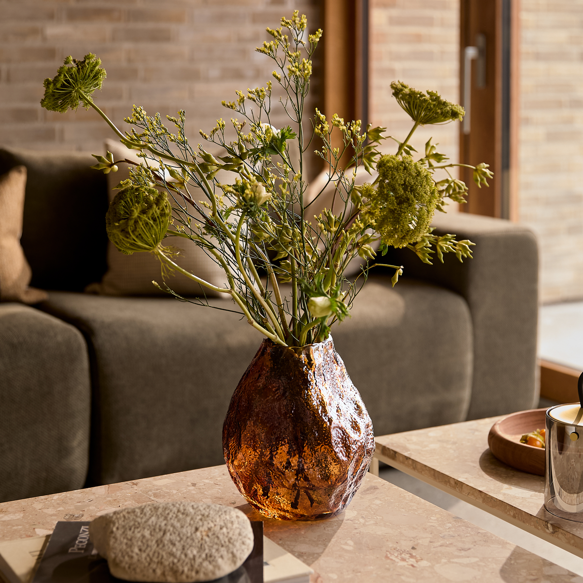 Ravfarvet, tekstureret glasvase med grønne grene på et sten-/marmor sofabord i en hyggelig stue med sofa i baggrunden.