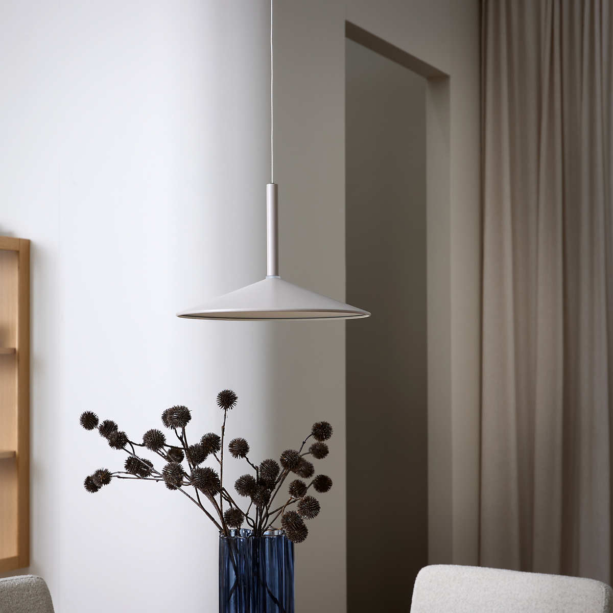 Hvid, konisk pendellampe over en blå glasvase med tørrede blomster i et lyst, minimalistisk rum med beige gardiner.